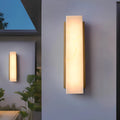 Modern rectangular outdoor wall light with gold frame and alabaster-style diffuser, softly glowing against a gray exterior wall