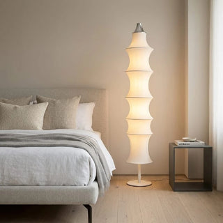 Celeste white silk modern floor lamp glowing warmly next to a bed in a minimalist bedroom