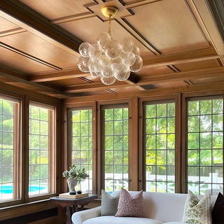 Round glass chandelier with gold chain hanging from wood coffered ceiling in bright living room.