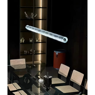 Illuminated glass tube pendant light suspended above a modern dining table with rattan chairs and glass cabinet in the background