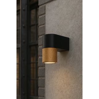 Modern outdoor wall sconce with a gold textured accent and black base mounted on a concrete exterior wall near large industrial-style windows