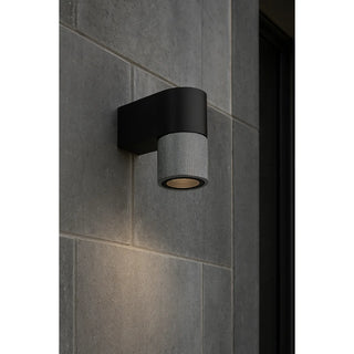 Gray textured modern wall sconce with a black base installed on a dark stone exterior wall, casting soft downward light