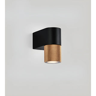 Close-up of a modern wall sconce with a single textured gold cylinder and a smooth black rectangular base on a light background